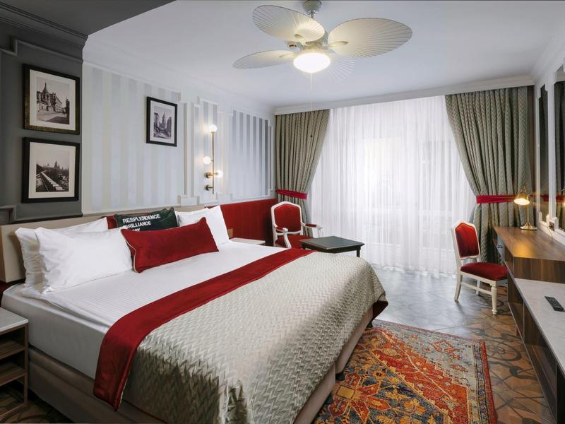 Modernes Hotelzimmer mit großem Bett, rot-weißer Bettwäsche, Teppich und Fenster mit Vorhängen.