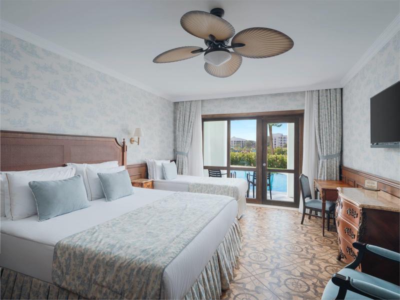 Helles Hotelzimmer mit zwei Doppelbetten, Deckenventilator und Balkon mit Blick aufs Wasser.