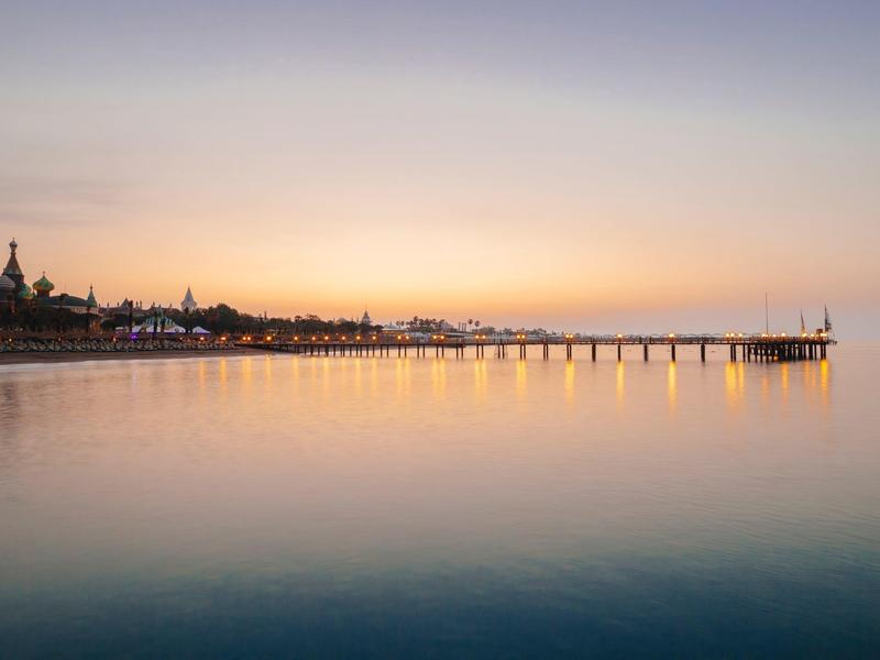 Langer Holzsteg im ruhigen Meer bei Sonnenuntergang, sanfte Farben am Himmel und im Wasser.