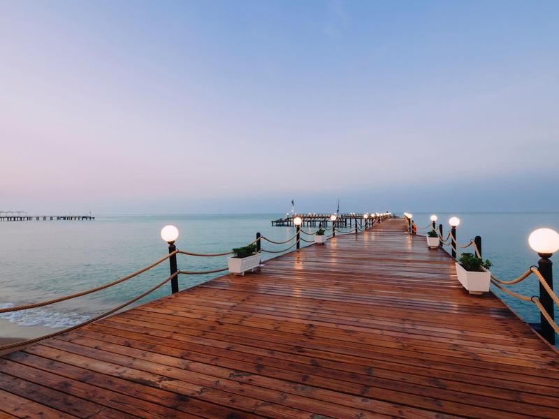 Langer Holzsteg mit Lampen an beiden Seiten, führt auf ruhiges, nebliges Meer hinaus.
