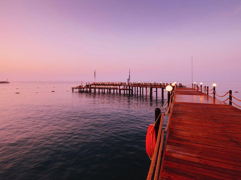 Holzsteg mit gelber Boje bei Sonnenuntergang über ruhigem Meer, weicher lila und oranger Himmel.