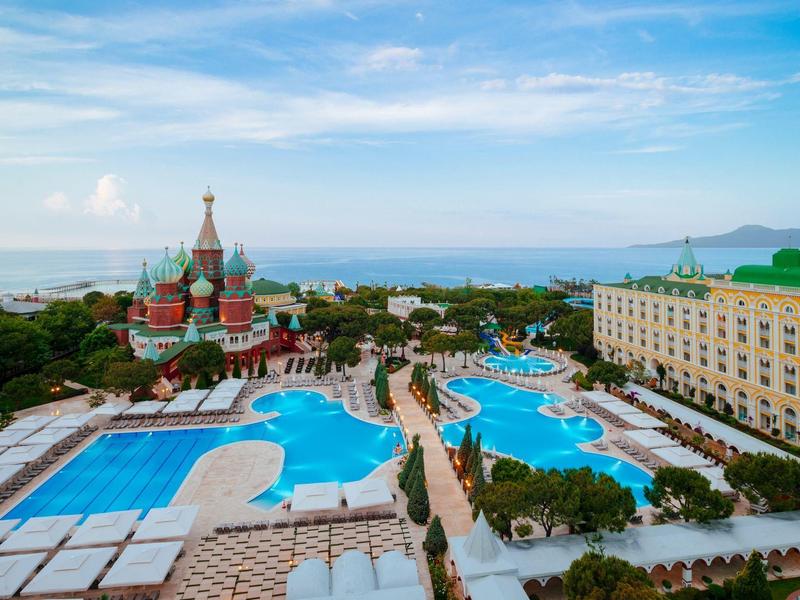Großer Hotelkomplex mit mehreren blauen Pools, Gartenflächen und einem Gebäude mit roten Türmen am Meer.