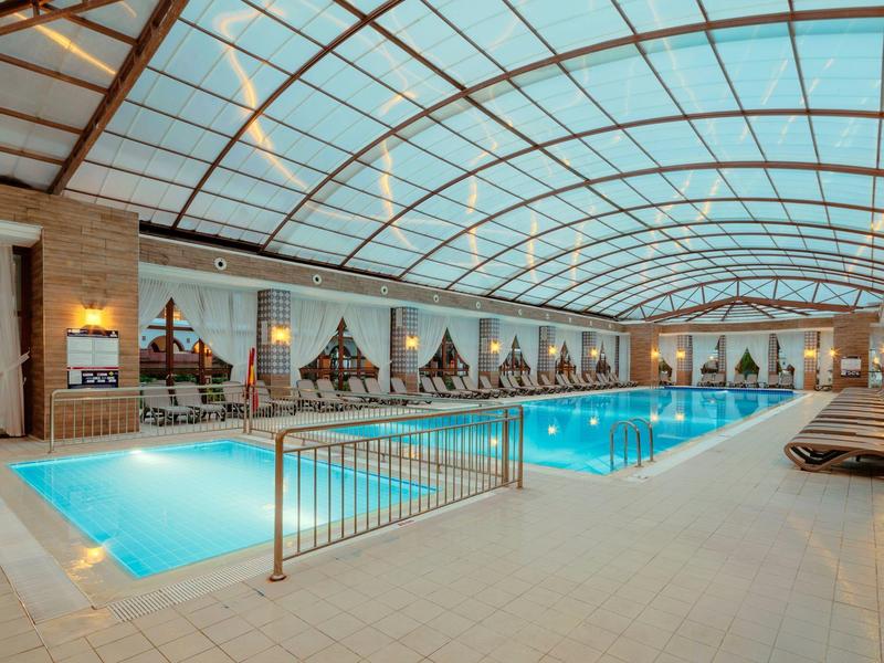 Großes Hallenbad mit gläsernem Gewölbedach, blauen Wasser und Liegestühlen am Rand.