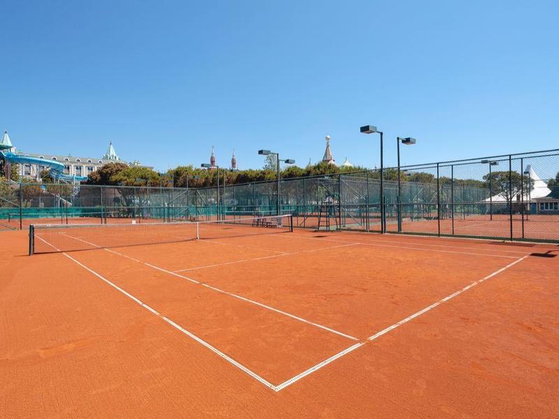Roter Sandplatz-Tennisplatz draußen, umgeben von Zaun und Lampen, unter klarem blauem Himmel.