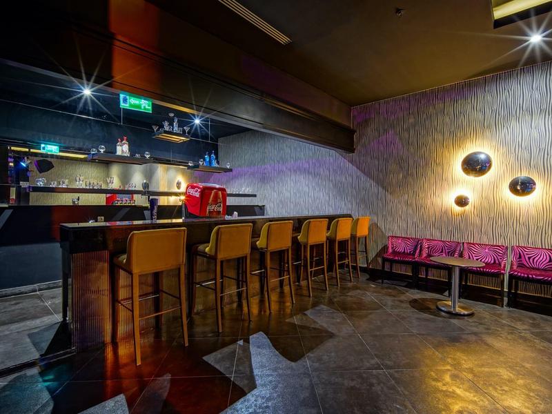 Bar d'hôtel moderne avec comptoir en bois, tabourets hauts et banquette violette devant un mur à motifs.