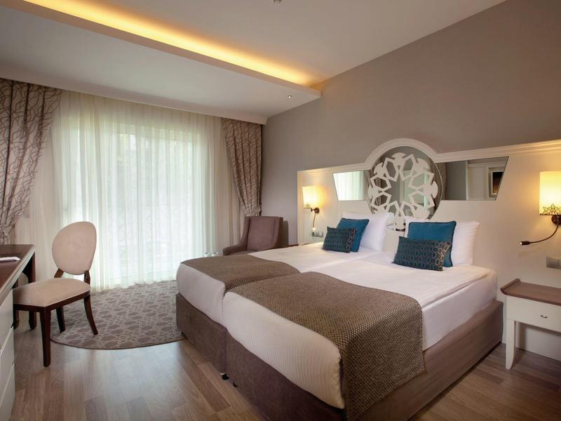 Chambre d'hôtel lumineuse avec lit double, deux tables de chevet, chaise et grande fenêtre avec rideaux.