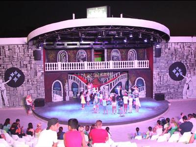 Spectacle sur scène avec un groupe de danseurs devant un décor de bâtiment historique, public assis au premier plan.