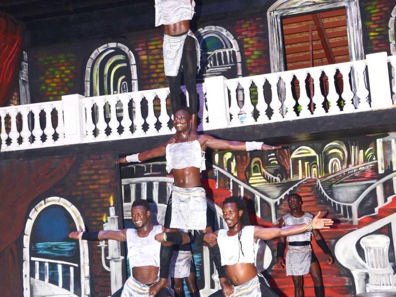 Artiesten vormen een menselijke piramide in een theater met kleurrijke geschilderde achtergronden en podiumverlichting.