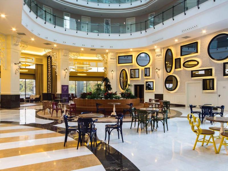 Ruime hotellobby met ronde tafels, stoelen en decoratieve muurspiegels onder een grote lichtkoepel.