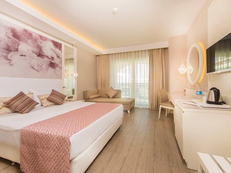 Lichte hotelkamer met tweepersoonsbed, bank, tv en groot raam met gordijnen.