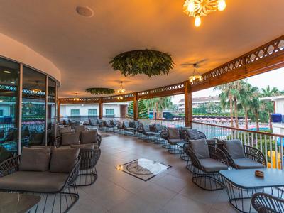 Terrazza coperta dell'hotel con sedie e tavoli neri, circondata da vetro con vista su palme.