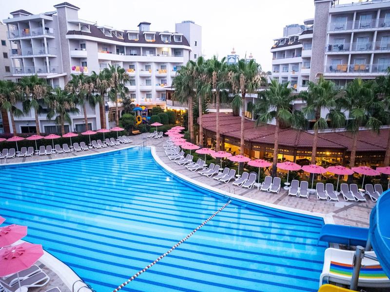 Duży basen na zewnątrz z leżakami i palmami przed wielopiętrowymi budynkami hotelowymi