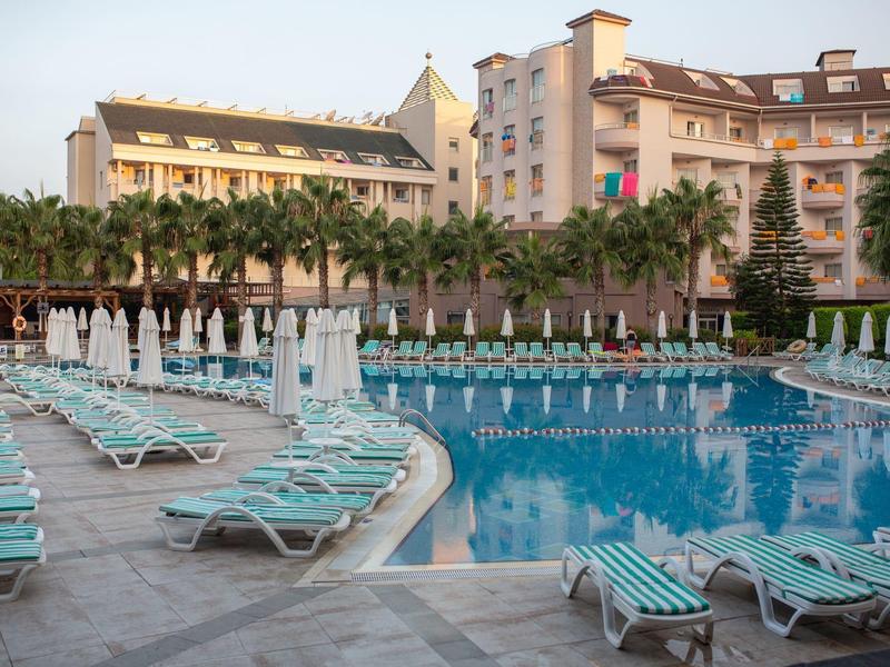 Duży basen hotelowy z leżakami i zamkniętymi parasolami, otoczony palmami i budynkami.