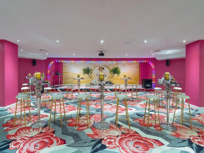 Nowoczesna sala eventowa z różowymi ścianami, wysokimi stołami i czerwonymi krzesłami.