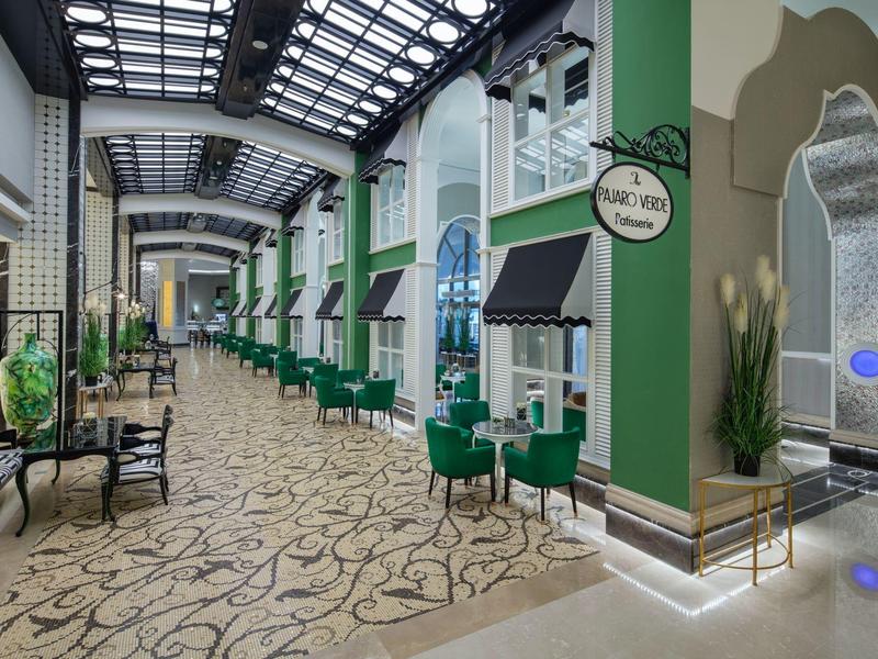 Przestronny korytarz hotelowy z zielonymi krzesłami, beżową wzorzystą podłogą i dużym szklanym dachem.