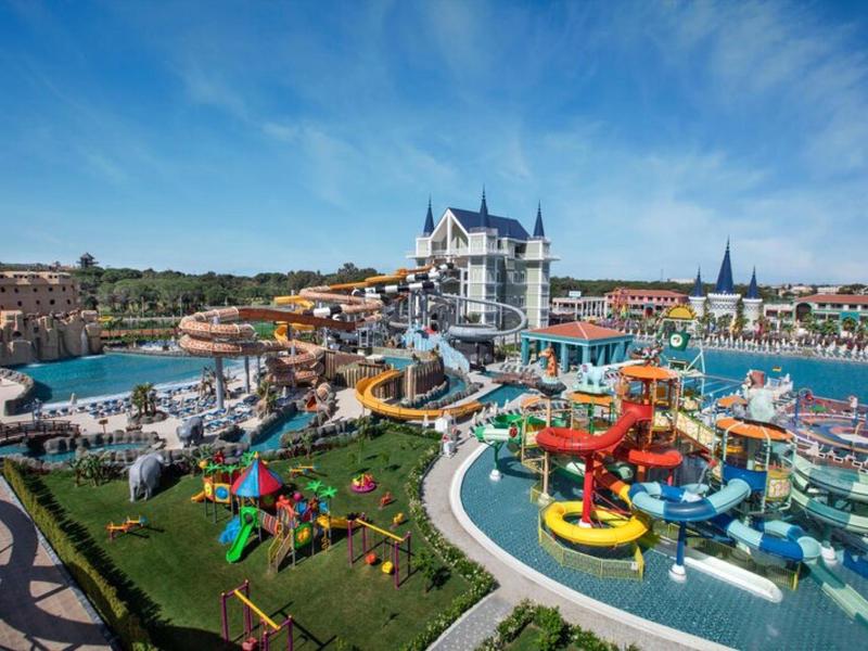 Park wodny z kolorowymi zjeżdżalniami, basenem i zamkiem w tle pod niebieskim niebem.