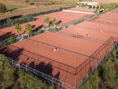 Mehrere rote Sandplätze für Tennis, umgeben von Zäunen und Bäumen in einer ländlichen Umgebung.