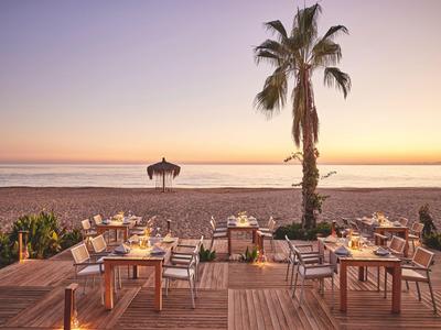 Strandrestaurant mit gedeckten Tischen, Palmen und Sonnenuntergang am Meer im Hintergrund.