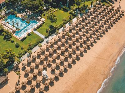 Vue aérienne d'une plage avec des rangées de parasols, un hôtel et une mer turquoise.