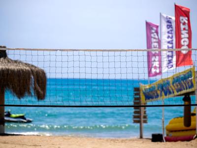Filet de beach-volley devant une mer bleue, plage de sable et drapeaux colorés en arrière-plan.