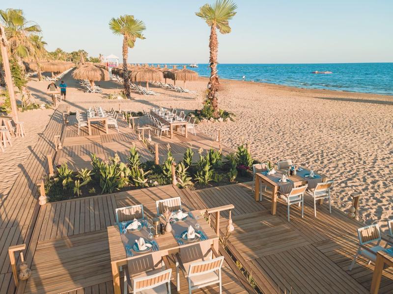 Strandrestaurant mit Holzboden, gedeckten Tischen und Palmen am Meer bei Sonnenuntergang.