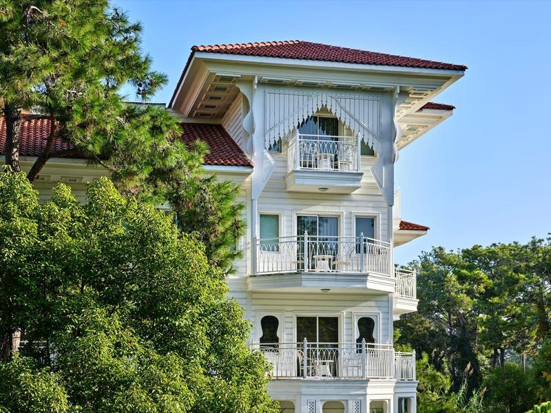 Weißes mehrstöckiges Hotelgebäude mit Balkonen und rotem Dach, umgeben von grünen Bäumen.