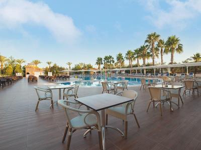 Große Terrasse mit Holzbohlen, Tischen und Stühlen neben einem Pool, Palmen im Hintergrund.