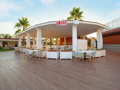 Modernes, offenes Restaurant mit weißem Runddach, Holzterrasse, Sitzgruppen und Palmen im Hintergrund.