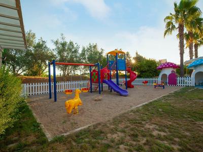 Kinderspielplatz mit bunten Rutschen, Schaukeln und Sandfläche, umgeben von Palmen und Gebäuden.