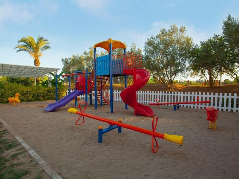 Plac zabaw dla dzieci z zjeżdżalnią, huśtawką i konstrukcją do wspinaczki otoczony drzewami i płotem.