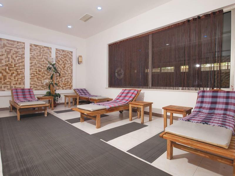 Jasne pomieszczenie relaksacyjne z drewnianymi leżakami, kraciastymi poduszkami i dużym oknem z żaluzjami.