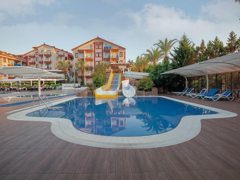 Basen hotelowy z zjeżdżalniami wodnymi, leżakami i parasolami, otoczony palmami i budynkami hotelu.