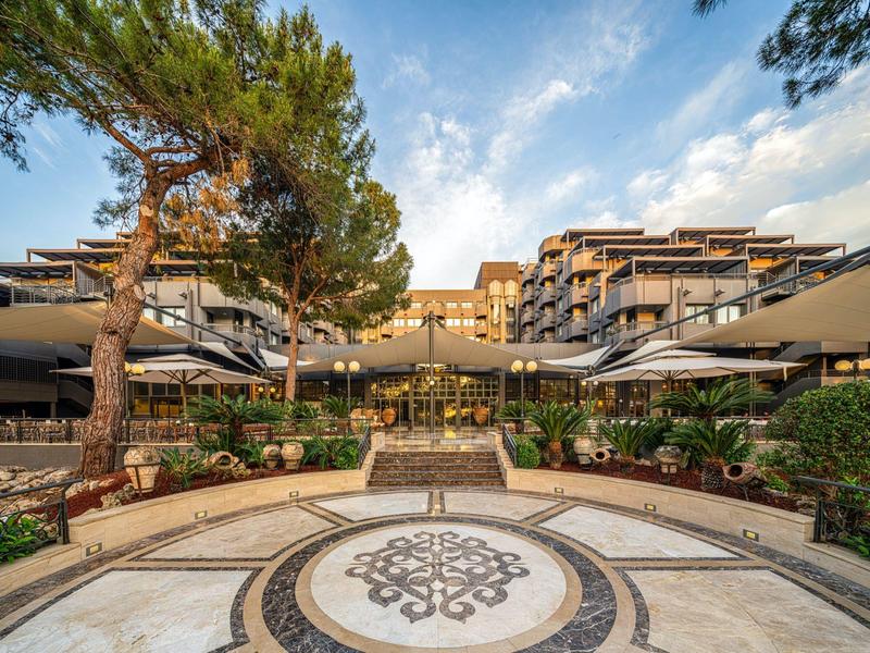 Modern hotel complex with multiple floors, surrounded by trees and a decorative mosaic in the foreground.