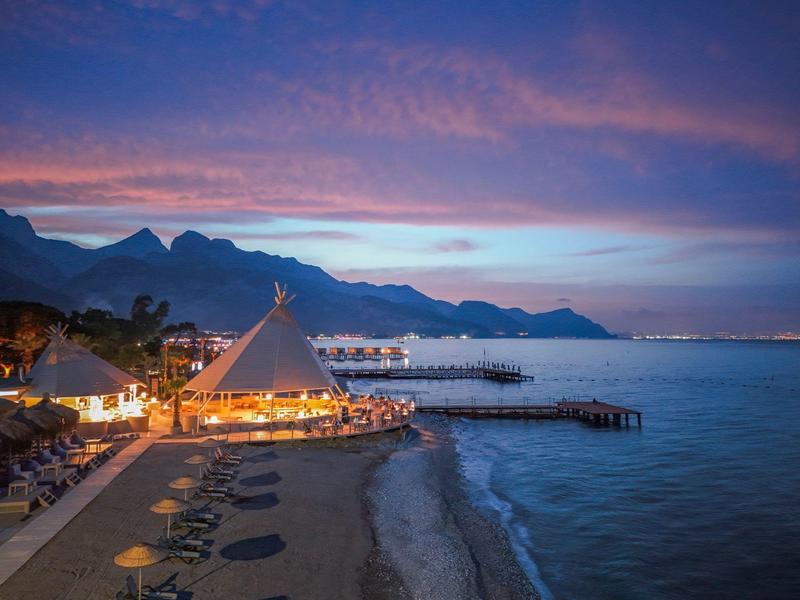 Beach with umbrellas, illuminated tent, and mountains at sunset by the sea.