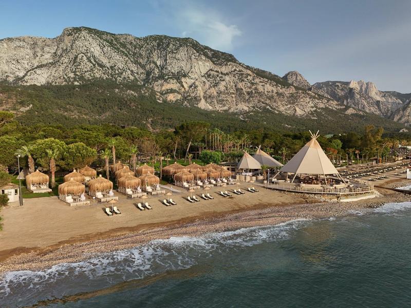 Beach with sun loungers and large tipi tents in front of a mountain landscape under clear sky.