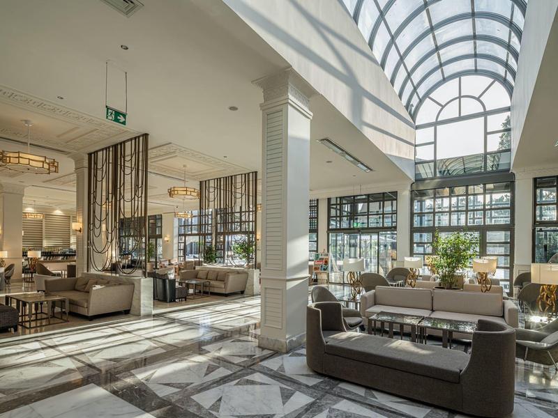 Spacious hotel lounge area with abundant natural light from a glass roof and elegant furnishings.