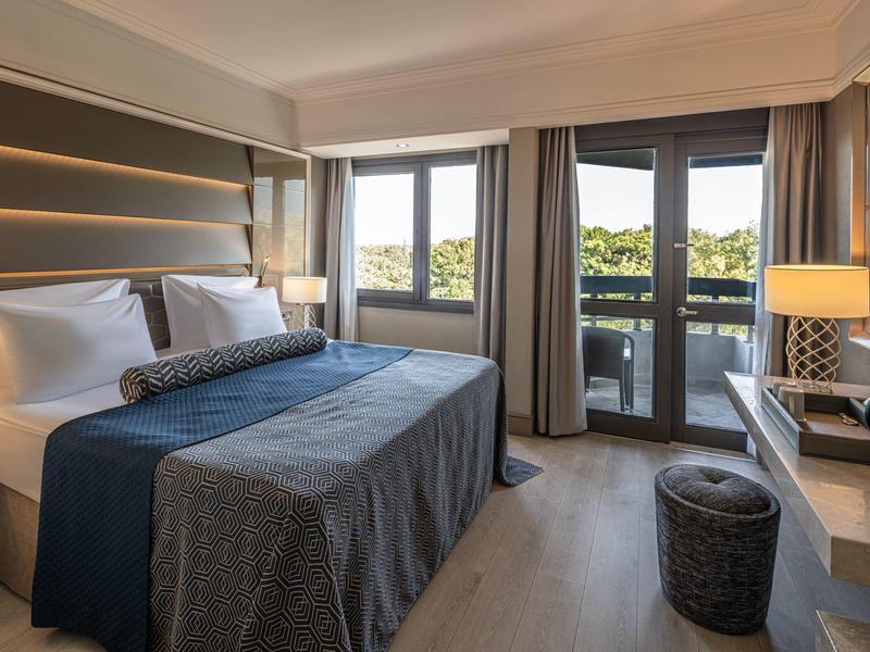 Modern hotel room with large bed, balcony, and green view.