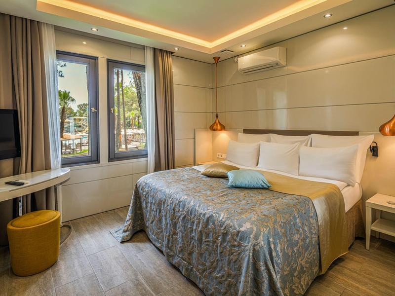 Modern hotel room with large bed, desk, window overlooking palm trees, and warm lighting.