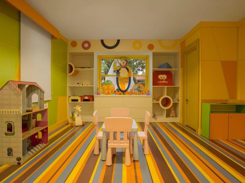 Children's playroom with colorful striped carpet, two chairs, a dollhouse, and shelves with toys.