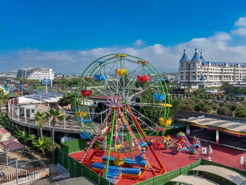 Colorful amusement park with Ferris wheel in front of historic hotel under blue sky