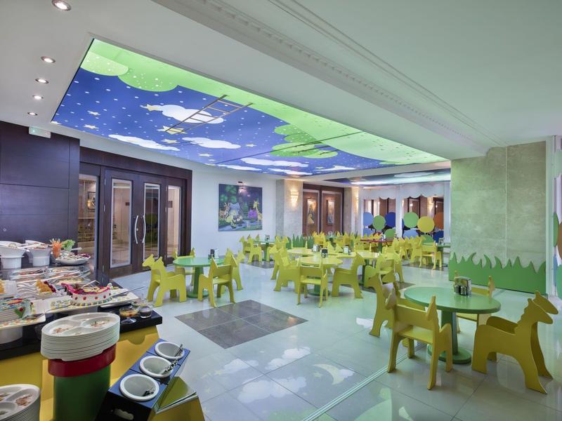 Kolorowa sala śniadaniowa z zielonymi krzesłami i dekoracją chmur na suficie.