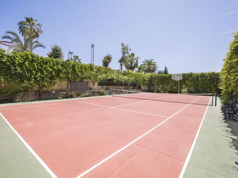 Pusty kort tenisowy z czerwonymi i zielonymi powierzchniami, otoczony palmami i kwitnącą zielenią.