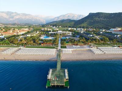 Strand mit Steg, dahinter grüner Park und Häuser vor Bergen unter blauem Himmel.