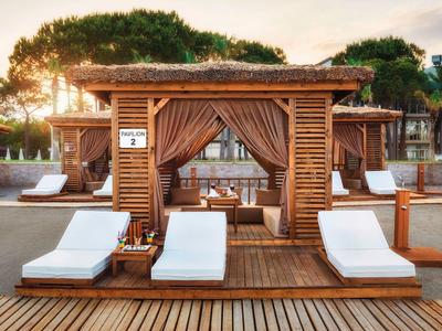 Zwei weiße Liegestühle vor einer Holzlounge mit Vorhängen und warmem Sonnenlicht.