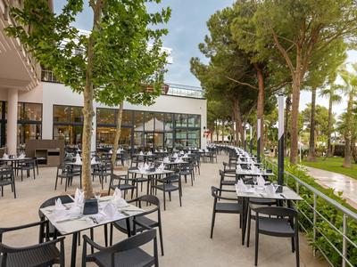 Große Außenterrasse mit vielen schwarzen Stühlen und weißen Tischen neben Bäumen und Grünfläche.