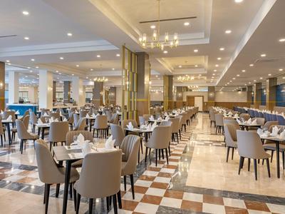 Großer heller Speisesaal mit vielen Tischen und beigen Stühlen, Marmorfliesen und moderner Beleuchtung.