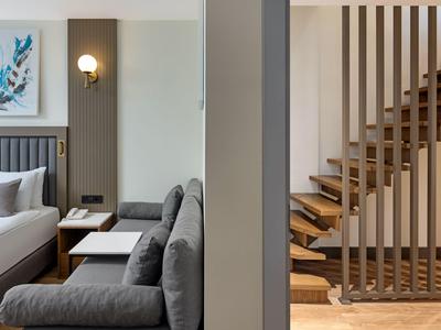 Camera d'hotel moderna con divano grigio, letto, scala in legno e design murale sobrio.