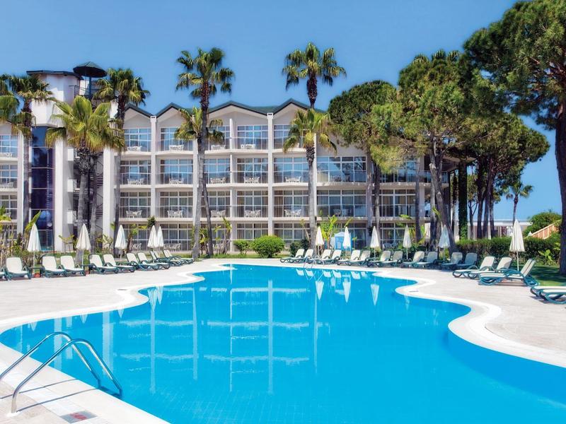 Großer, geschwungener Pool mit blauer Wasseroberfläche vor Hotel mit Glasfassade und Palmen.