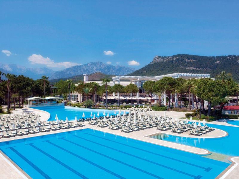 Großer Außenpool mit Liegestühlen, Palmen und Bergen im Hintergrund unter blauem Himmel.