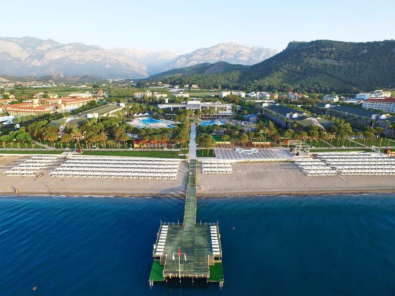 Strand mit Steg, dahinter grüner Park und Häuser vor Bergen unter blauem Himmel.